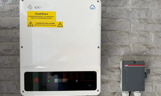En vit GoodWe växelriktare 10kW monterad på en grå stenvägg. Bredvid sitter en grå ABB-box.