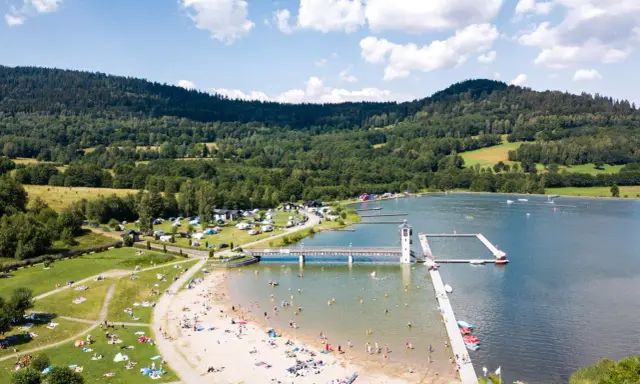 Słoneczny dzień nad zalewem w Starej Morawie. Ludzie odpoczywają na plaży i w wodzie, a w tle widać zielone wzgórza.