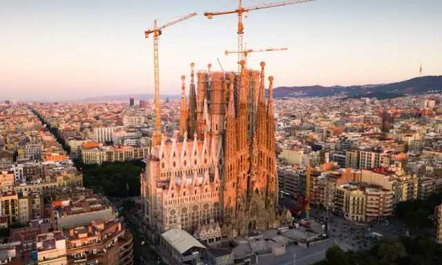 Widok z lotu ptaka na Sagrada Familia w Barcelonie, wciąż w budowie, z dźwigami górującymi nad miastem. Słońce oświetla fasadę, tworząc ciepłą poświatę, jakby przygotowując się do wieczornych mszy.