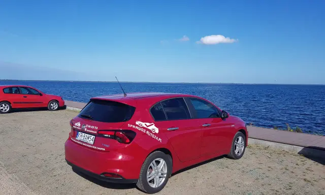 Czerwony fiat tipo sedan z napisem "SprawdzAuto24.pl" stoi nad błękitnym jeziorem w słoneczny dzień.