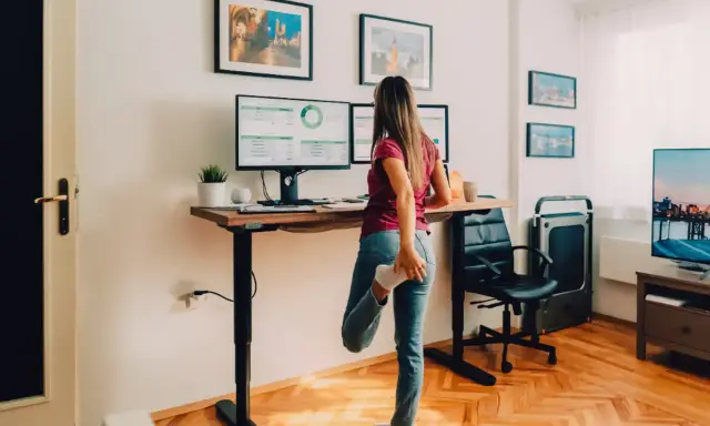 Femme s'étirant près de son bureau réglable en hauteur, pour un confort au bureau optimal. Deux écrans affichent des graphiques.