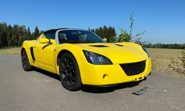 Żółty Opel Speedster na tle lasu i pól. Słońce pięknie podkreśla jego sportową sylwetkę.