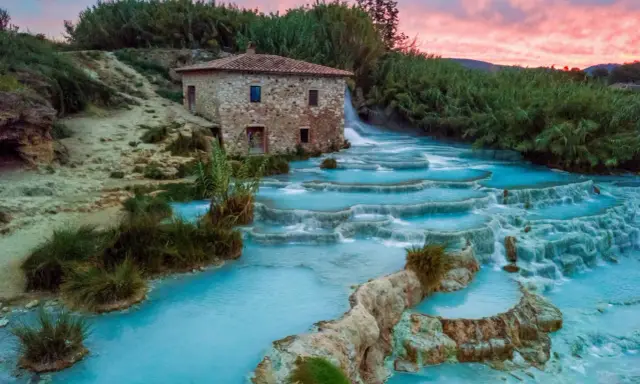 Cascate termali turchesi in Toscana, con un antico mulino in pietra e un cielo rosa al tramonto. Curiosità sulla Toscana: un luogo magico.