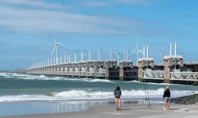 Dwie osoby stoją na plaży, obserwując falujące morze i imponującą zaporę z turbinami wiatrowymi. Takie morze jest w Holandii, chronione przez innowacyjne konstrukcje.