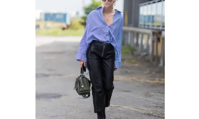 Donna con camicia a righe blu e bianche, occhiali da sole e un elegante outfit pantaloni ecopelle neri con zip frontale. Porta una borsa verde oliva.