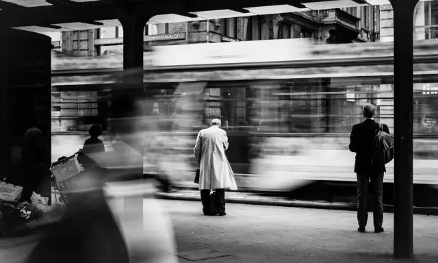 Street photography esempi: un tram sfreccia, lasciando figure sfocate in attesa alla fermata.