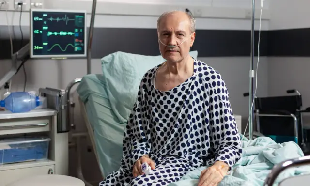Starszy mężczyzna w szpitalnym łóżku, z tlenem w nosie, walczy z ostrą białaczką szpikową. Monitor pokazuje jego parametry życiowe.