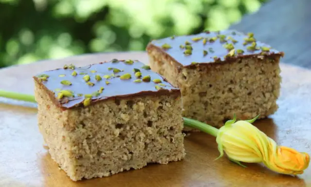 Zwei Stücke saftiger Zucchini Kuchen mit Schokoladenglasur und gehackten Pistazien, daneben eine Zucchiniblüte.
