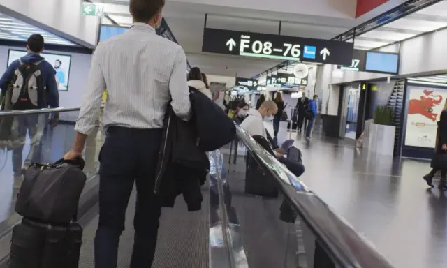 Resenärer på rullband på flygplatsen, skyltar visar vägen till gate F08-76. En man med resväskor på väg mot centrum.