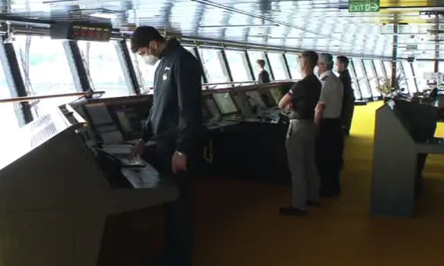 Equipaggio sulla plancia nave da crociera, con strumenti di navigazione e grandi finestre.