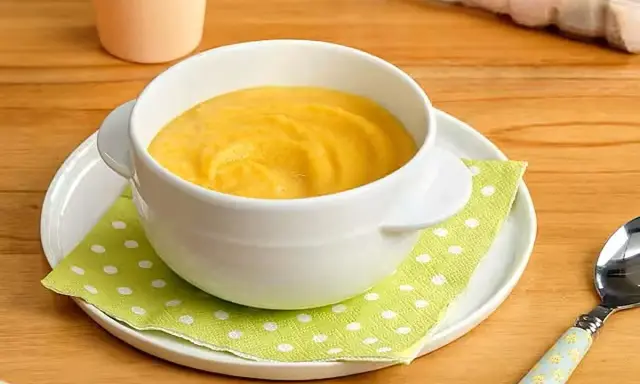 Un tazón blanco con puré de pescado y arroz para bebé, servido sobre un plato blanco y una servilleta verde con puntos blancos.