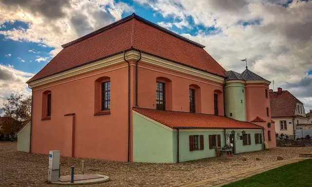 Tykocin co zobaczyć: zabytkowa Wielka Synagoga z różową fasadą i zielonym aneksem, na brukowanym placu pod burzliwym niebem.