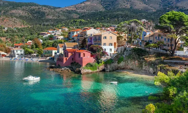 Case colorate affacciate su acque cristalline, un sogno di spiagge Grecia. Barche a vela e colline verdi completano il quadro.