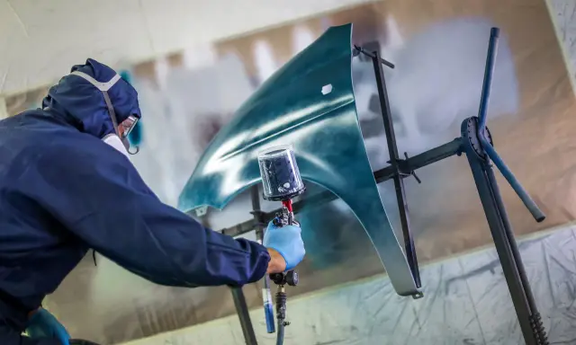 Pracownik w kombinezonie ochronnym wykonuje lakierowanie wybiórcze błotnika samochodu, używając pistoletu natryskowego.