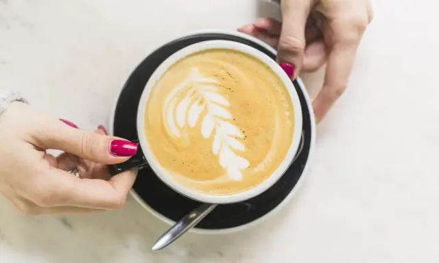 Une main tient une tasse de café latte avec un dessin de feuille. Un guide pour comment faire un latte parfait.