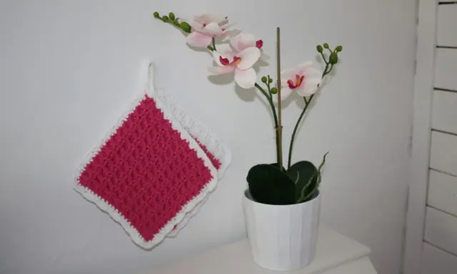 Två tjocka, virkade grytlappar i rosa med vit kant hänger på en vit vägg bredvid en orkidé i kruka.