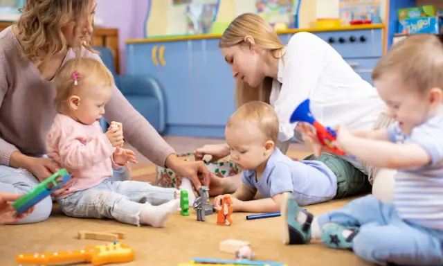 Opiekunki bawią się z dziećmi zabawkami na dywanie w żłobku.