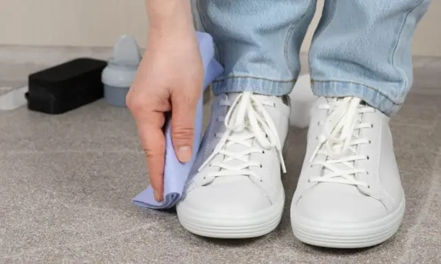 Persona limpiando zapatillas blancas con un paño azul. A su lado, un cepillo y un bote de limpiador.