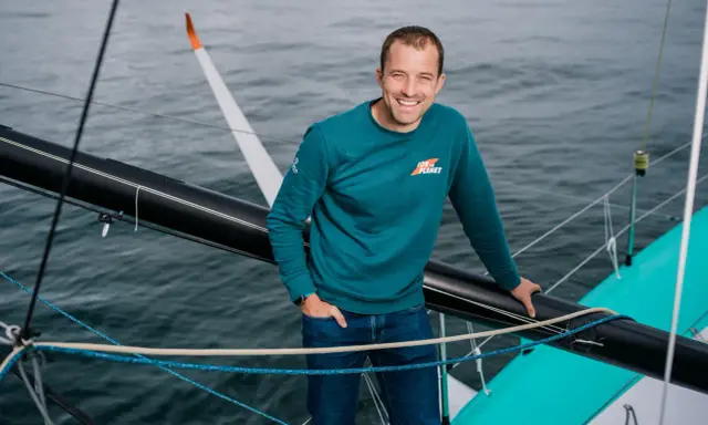 Sam Goodchild, souriant, sur un voilier, portant un sweat "For the Planet".