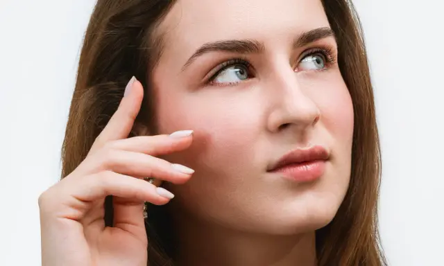 Une jeune femme aux yeux bleus regarde au loin, sa main effleurant sa joue. Ses cils, sublimés par un **tatouage cils**, accentuent son regard captivant.