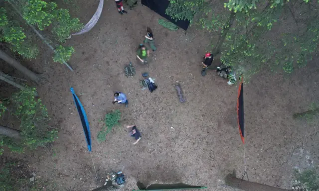 Ludzie rozbijają obóz w lesie, stosując zasady "leave no trace". Widać hamaki, plecaki i namiot.