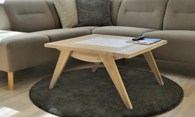Ett smart soffbord i ljust trä med en vit inläggning, placerat på en rund, mörk matta framför en beige hörnsoffa.