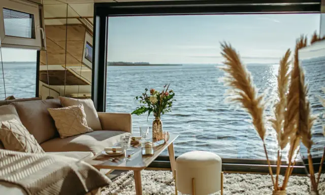Przytulny salon z widokiem na wodę, idealny na nietypowe noclegi w Polsce. Sofa, stolik z kwiatami i puszysty dywan tworzą atmosferę relaksu.