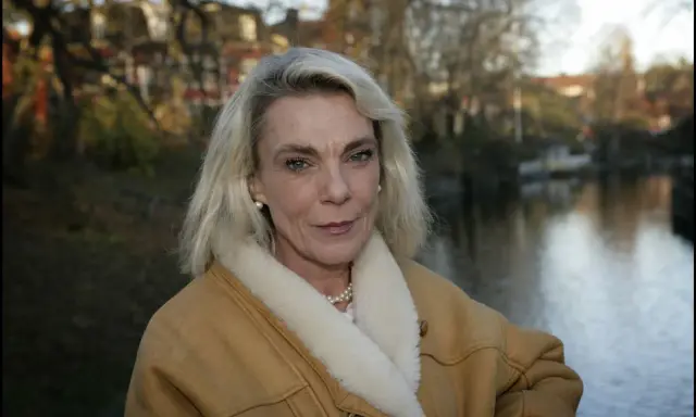 Pia Degermark idag, med sitt blonda hår och en varm fårskinnsjacka, står vid en vattensamling med träd och hus i bakgrunden.