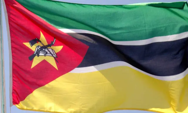 Flaga Mozambiku: zielono-czarno-białe pasy, czerwony trójkąt z żółtą gwiazdą, karabin AK-47 i książka.