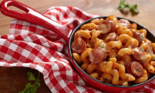Pyszne spaghetti z kiełbasą w sosie pomidorowym, podane w czerwonej patelni, obok czerwono-białej kratkowanej serwetki.