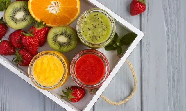 Bandeja con fresas, rodajas de kiwi, naranja y tres tarros de **puré de frutas** de colores vibrantes: verde, amarillo y rojo.