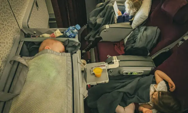 Lot z niemowlakiem i dwójką śpiących dzieci w samolocie. Niemowlę śpi w gondoli, starsze dzieci odpoczywają pod kocami.