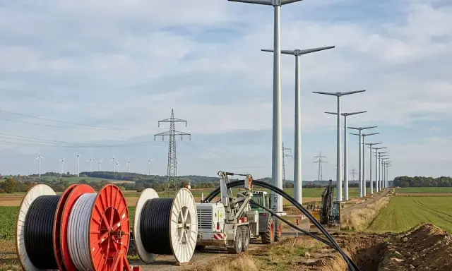 Kabeltrommeln und Bagger im Feld, während neue Windräder aufgestellt werden. Die Energiewende Herausforderungen bei der Infrastruktur sind sichtbar.