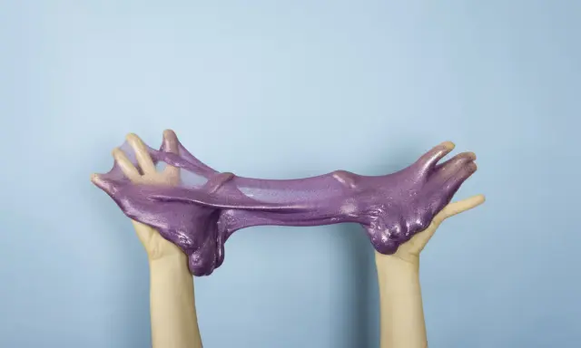 Manos estirando un brillante slime casero morado con purpurina sobre fondo azul.