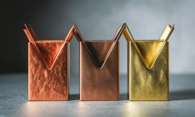 Trois porte-stylos aux formes géométriques, aux teintes cuivrées et laiton couleur, sur un fond gris.