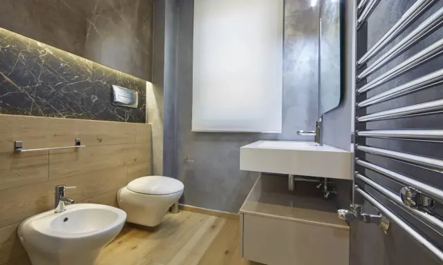 Bagno moderno con bidet, WC sospeso, lavabo e scaldasalviette cromato. L'illuminazione a LED sotto il marmo crea un'atmosfera elegante, rispettando l'altezza minima soffitto.