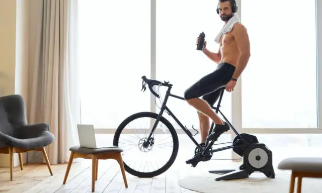 Mężczyzna w słuchawkach pije wodę, ćwicząc na rowerze stacjonarnym przy dobrej muzyce na rower.
