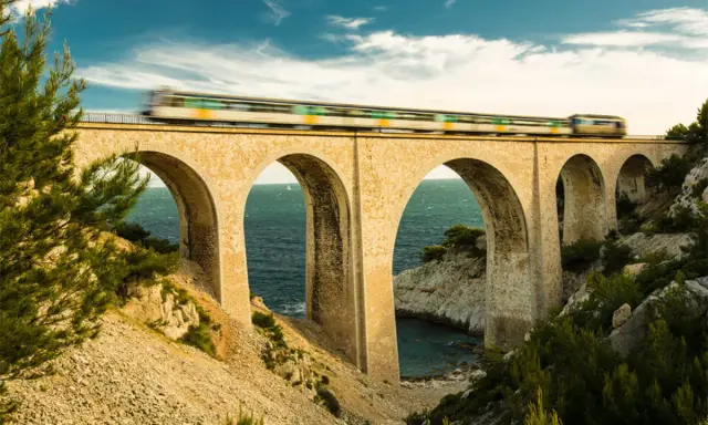 Ein Zug rast über eine Bogenbrücke, die Teil des Bahnnetz Frankreichs ist. Das Meer glitzert unter den Bögen.