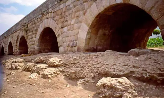Antiguo puente de piedra con arcos, que es un puente de gran tamaño y robustez, con ruinas a sus pies.