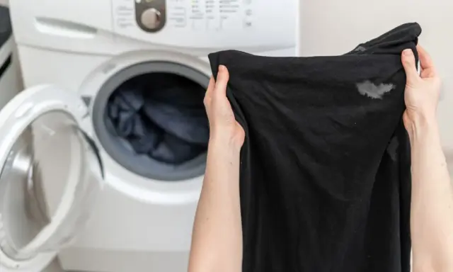 Manos sostienen ropa negra con manchas blancas, cerca de una lavadora abierta. ¿Por qué la ropa negra sale con manchas blancas?