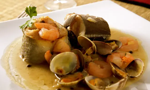 Delicioso abadejo a la donostiarra con gambas y almejas en una salsa dorada, adornado con perejil fresco.