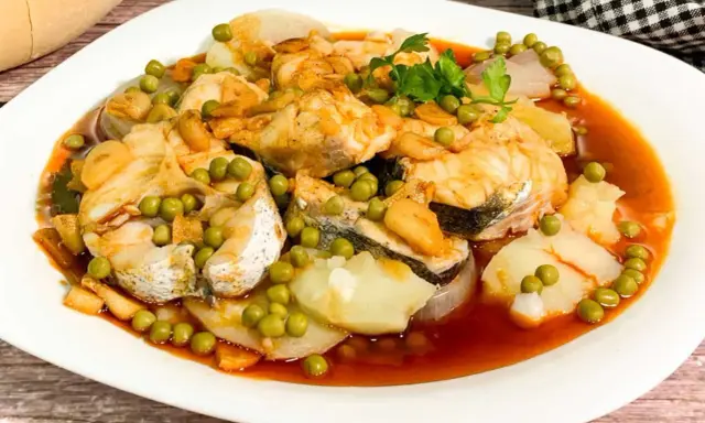 Merluza a la gallega con patatas, guisantes y ajo frito en una salsa rojiza.
