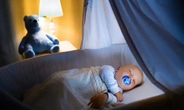 Bambino dorme sereno nella culla, con ciuccio, sotto una coperta calda. Un orsacchiotto veglia, simbolo di addormentamento autonomo.