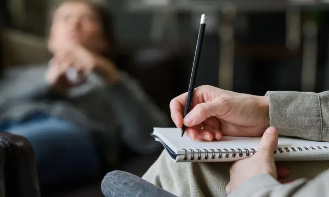 Psycholog pomaga zrozumieć siebie i swoje problemy. Na zdjęciu widać rękę terapeuty piszącą w notatniku, podczas gdy pacjent leży na kanapie.