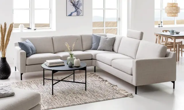 Ljusbeige hörnsoffor i ett modernt vardagsrum med ett runt soffbord, en matta och stora fönster.