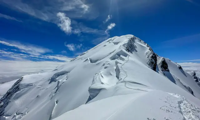 Śnieżna trasa na Mont Blanc, z widocznymi szczelinami lodowcowymi i śladami wspinaczy. Błękitne niebo z chmurami.