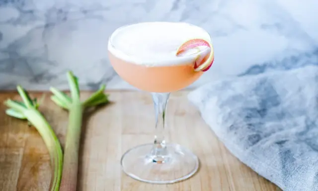 En rosa rabarber sour-drink med skum och en rabarberdekoration. Två stjälkar rabarber ligger bredvid.
