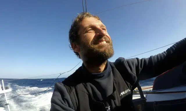 Un homme de mer souriant, le visage tourné vers le ciel bleu, navigue sur un voilier. L'océan agité et le soleil radieux témoignent de son aventure.