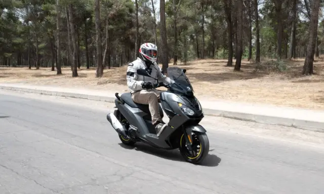 Hombre en casco y chaqueta conduce un triciclo de motor gris por una carretera rodeada de pinos.