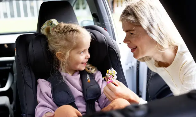 Uśmiechnięta mama daje córeczce w foteliku samochodowym bukiecik kwiatów. To miły test plus dla małej pasażerki.